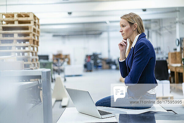Thoughtful businesswoman with hand on chin sitting on desk in factory