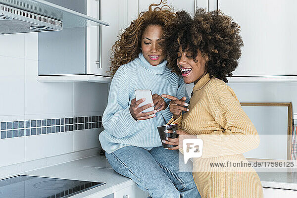 Young woman sharing mobile phone with friend in kitchen