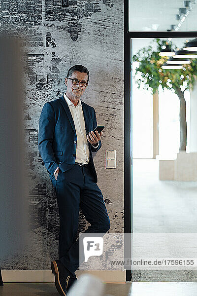 Businessman with hand in pocket holding smart phone leaning on wall at office