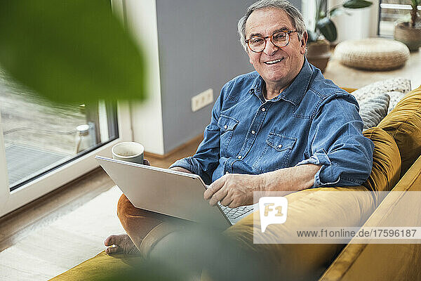 Smiling senior man with laptop and coffee mug sitting on sofa at home