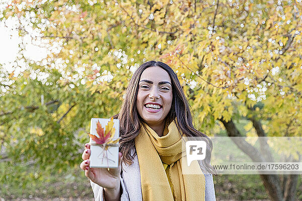 Fröhliche junge Frau hält kleine Geschenkbox mit Herbstblatt im Park
