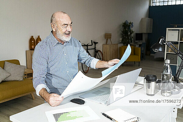 Smiling senior freelancer reading documents sitting in home office