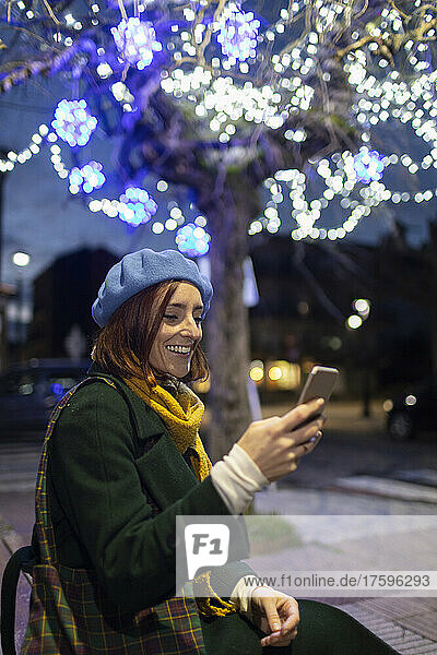 Happy woman text messaging through smart phone sitting in front of illuminated tree at night