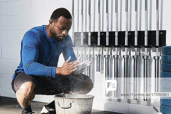 Athlete applying chalk powder crouching near bucket in gym