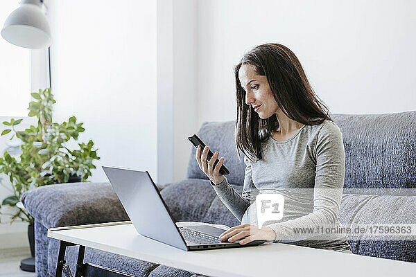 Pregnant woman with smart phone working on laptop at home office