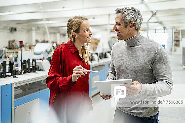 Happy blond businesswoman sharing ideas with colleague holding tablet PC in factory