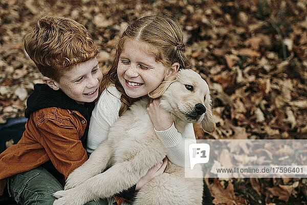 Happy girl embracing golden retriever puppy by brother on autumn leaves in forest