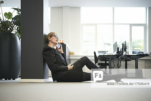 Geschäftsfrau spricht mit Smartphone und sitzt im Büro auf dem Boden