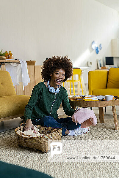 Happy woman with curly hair folding clothes sitting on carpet in living room