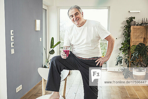 Smiling senior man holding glass of juice standing with hand on hip at home