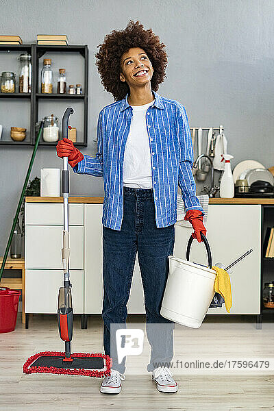 Thoughtful young Afro woman holding cleaning bucket and mop in kitchen