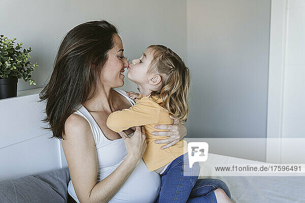 Cute girl kissing pregnant mother's nose on bed at home