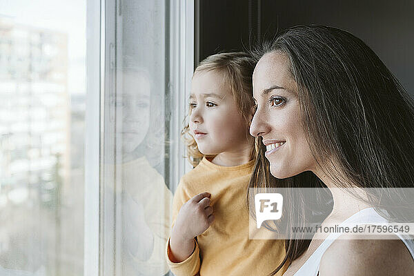 Smiling woman with daughter looking through window at home