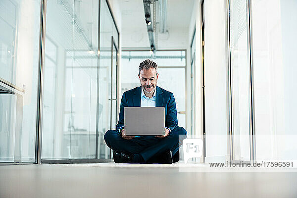 Businessman sitting cross-legged using laptop in office corridor
