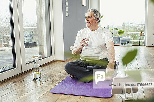 Smiling senior man with drinking glass sitting cross-legged on exercise mat at home