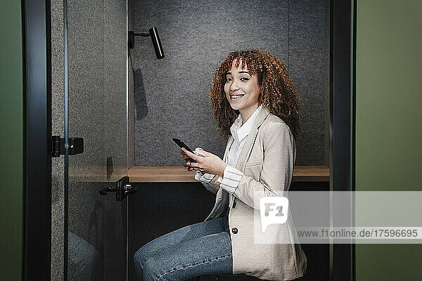 Smiling young businesswoman with smart phone sitting at desk in cabin