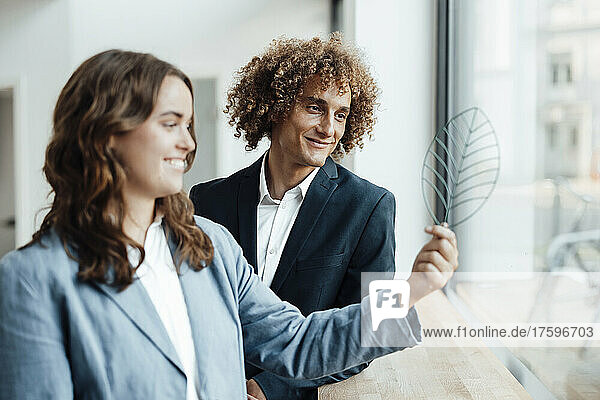 Smiling businessman and businesswoman looking at leaf model by glass window in office