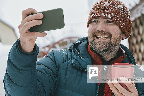 Smiling man holding coffee mug taking selfie through mobile phone in winter