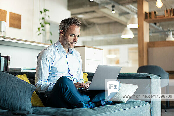 Geschäftsmann sitzt mit gekreuzten Beinen auf Sofa im Büro und benutzt Laptop