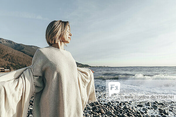 Blond woman with eyes closed holding blanket at beach