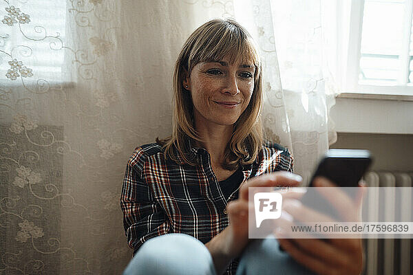 Smiling woman using smart phone by window at home