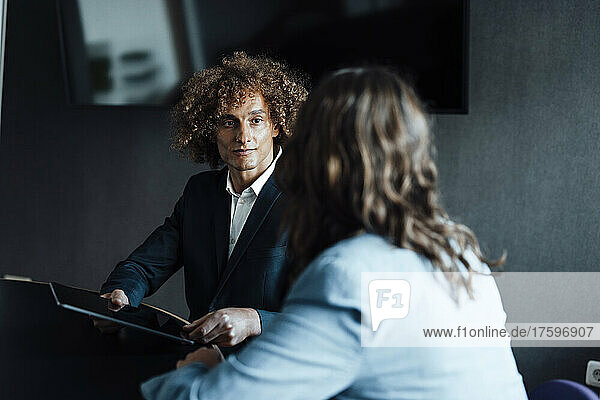 Curly haired businessman holding tablet PC looking at colleague discussing in coworking office