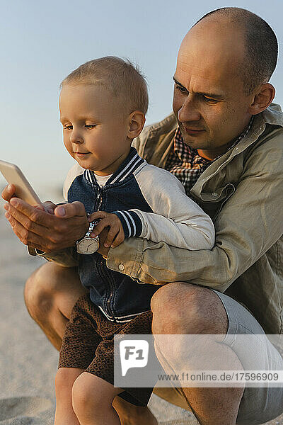 Netter Junge mit Vater  der sein Smartphone am Strand bei Sonnenuntergang benutzt