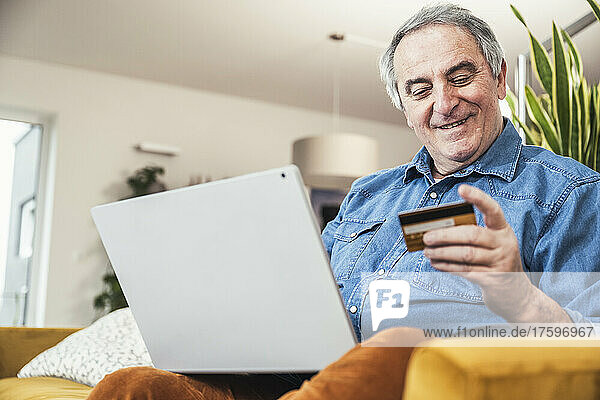 Smiling senior man with laptop looking at credit card in living room at home