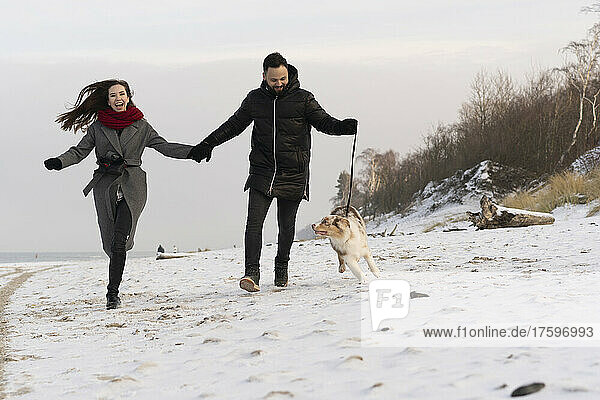 Happy young couple running with dog on snow at sunset