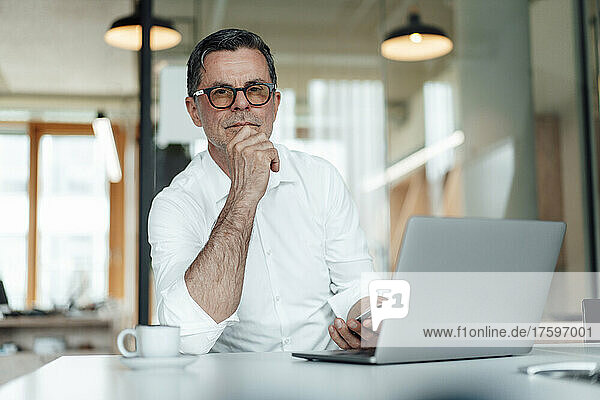 Confident businessman with hand on chin sitting with laptop on table at office