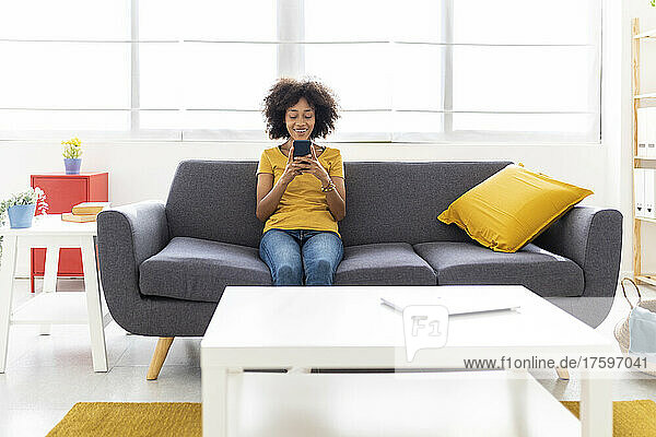 Smiling young woman using smart phone sitting on sofa in living room at home