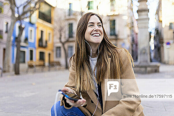 Happy woman talking on smart phone through in-ear headphones at city street