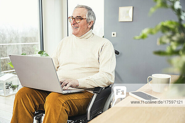 Smiling disabled man wearing eyeglasses sitting with laptop on wheelchair