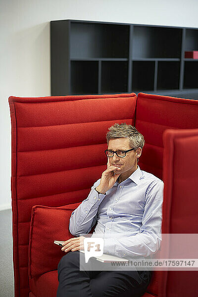 Thoughtful businessman with laptop sitting in red chair at office