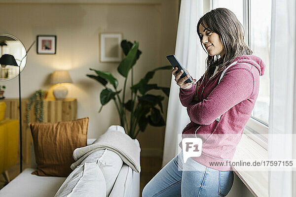 Woman text messaging through smart phone by window at home