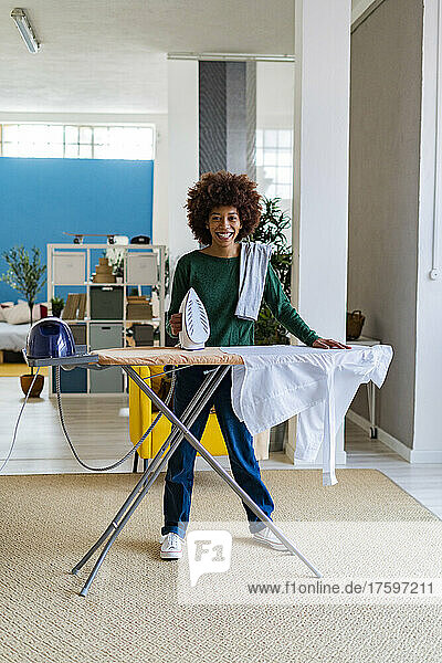Smiling young Afro woman with shirt on board holding iron in living room