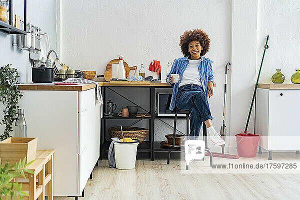 Smiling young Afro woman sitting on stool leaning on kitchen counter relaxing at home