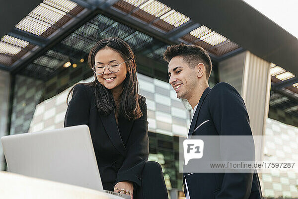 Smiling business colleagues working on laptop in front of office building