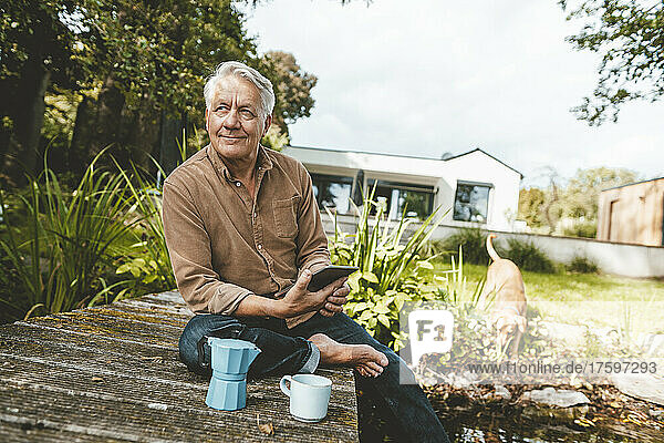 Lächelnder älterer Mann mit Tablet-PC sitzt auf der Anlegestelle im Hinterhof