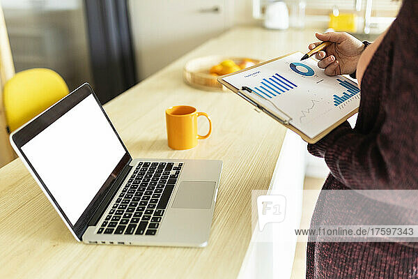 Eine berufstätige Frau überprüft Diagramme und hält die Zwischenablage am Laptop auf dem heimischen Tisch