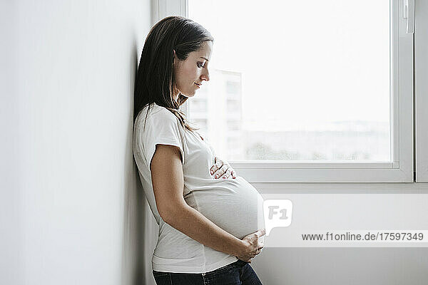 Schwangere Frau lehnt zu Hause an der Wand am Fenster
