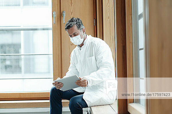 Scientist with protective face mask using tablet PC sitting near window at clinic