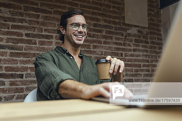 Smiling businessman with disposable coffee cup using laptop in office