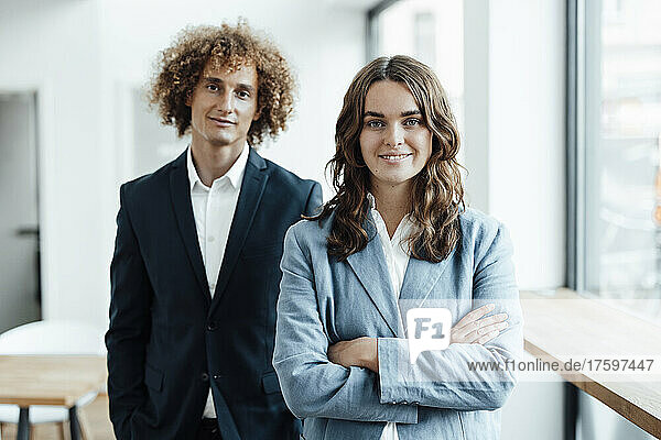 Smiling young businesswoman with arms crossed standing in front of colleague in office