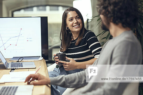 Lächelnde Geschäftsfrau hält Kaffeetasse in der Hand und unterhält sich mit einem Geschäftsmann  der am Konferenztisch sitzt