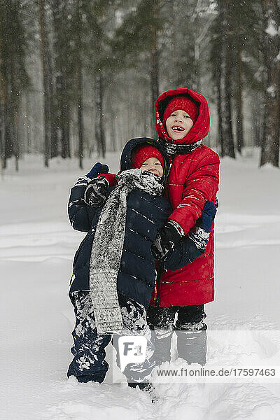 Smiling boy embracing brother standing on snow in forest