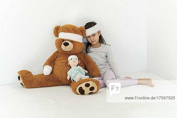 Bandage on girl with teddy bear and doll sitting in front of white wall