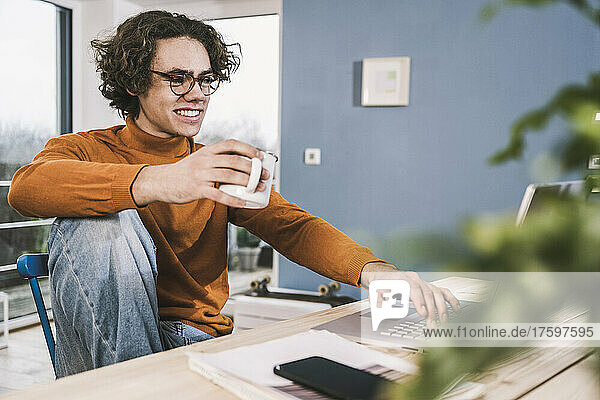 Happy young man holding coffee cup using laptop at home
