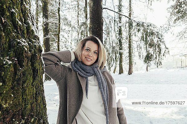 Smiling woman standing with hands behind head by tree in winter forest