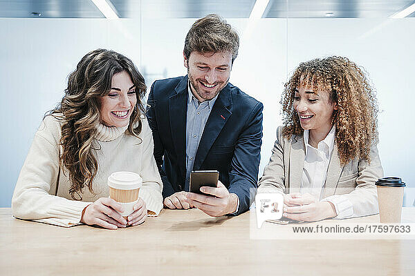 Happy businessman sharing smart phone with businesswomen sitting at desk